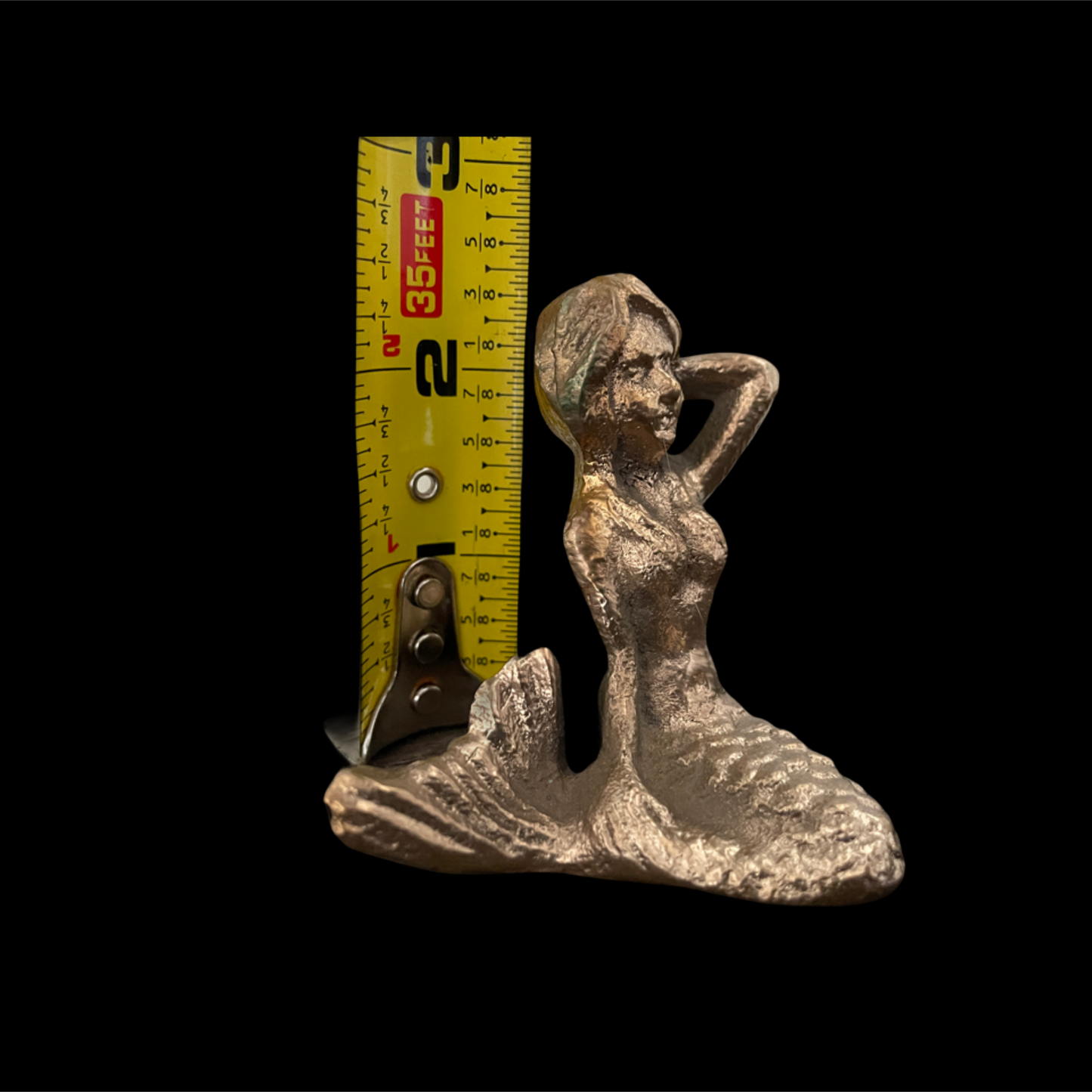 Metallic mermaid figurine next to a measuring tape on a black background